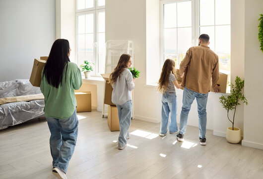 Family parents children moving boxes into home. Back view shows them near bright windows with cartons, exploring the new space. Soft daylight fills the room. Bright family move concept image