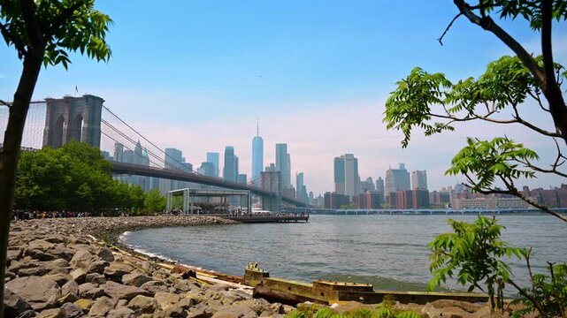 Rocky waterfront of the East River with crowds of people in the backdrop. Low angle view at the Brooklyn Bridge and famous Manhattan skyscraper skyline at daytime.