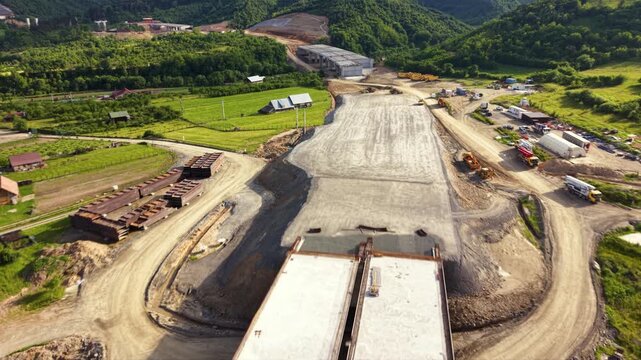 Aerial views of a major highway viaduct under construction across a broad mountain valley, showing completed concrete bridge deck sections, active earthworks, cranes and a nearby road interchange besi