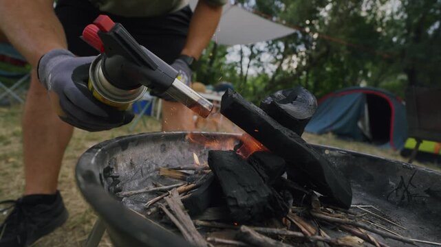 Male camper using a handheld torch to ignite charcoal and kindling in a fire pit surrounded by camping gear and tents in a natural outdoor setting