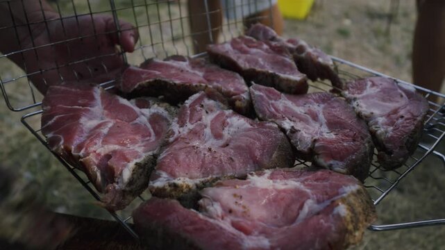 Close-up of hand placing marinated meat slices on a grill basket outdoors, showcasing the preparation of a barbecue meal in a grassy area with blurred background elements