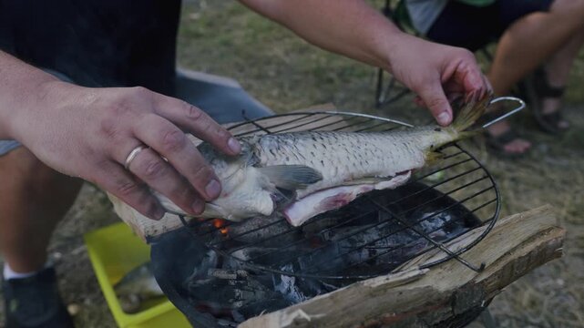 Person preparing fish on a grill over charcoal, showcasing the process of cooking outdoors with a yellow container and grassy ground in the background