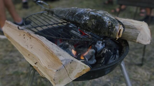 Whole fish grilling on a charcoal barbecue with flames and wooden logs, surrounded by a natural outdoor setting with blurred figures in the background