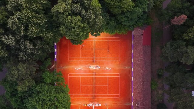 Top-down aerial view of a red clay tennis court surrounded by dense green trees, with crisp white court markings and warm sunlight creating a strong geometric sports pattern.