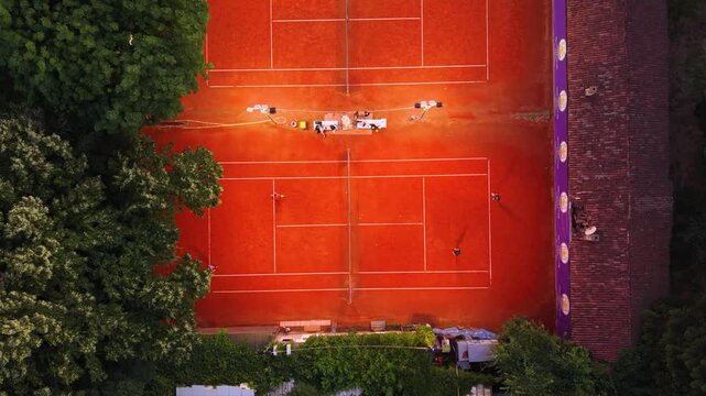 Top-down aerial view of a red clay tennis court surrounded by dense green trees, with crisp white court markings and warm sunlight creating a strong geometric sports pattern.