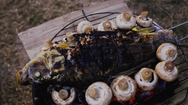 Grilled fish with mushrooms on a barbecue grill, showcasing the cooking process with visible flames and smoke in an outdoor setting on a wooden surface