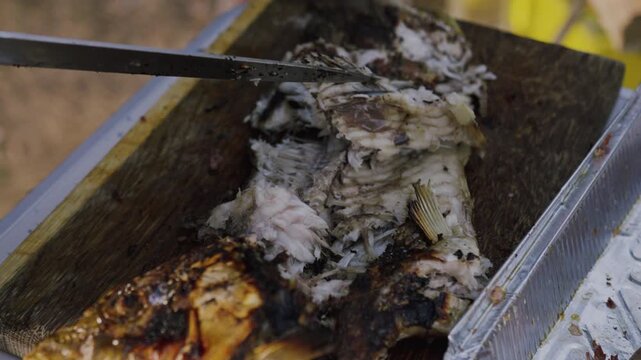 Grilled fish being sliced on a wooden cutting board with a knife, showcasing the texture and details of the fish in an outdoor setting with natural lighting