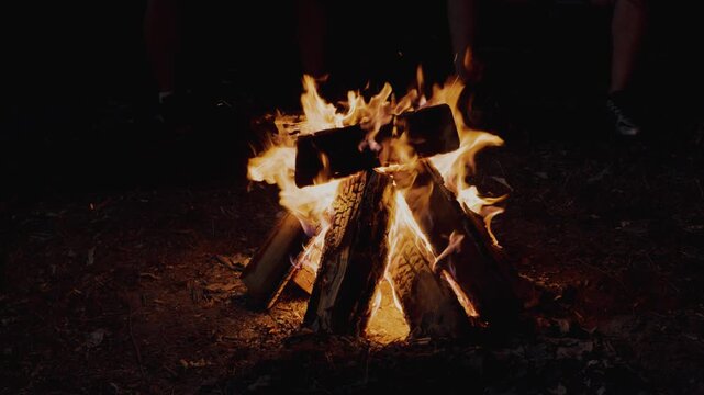 Campfire flames dance around stacked logs as sparks fly into the night sky, creating a warm glow in the dark forest setting with silhouettes of people nearby