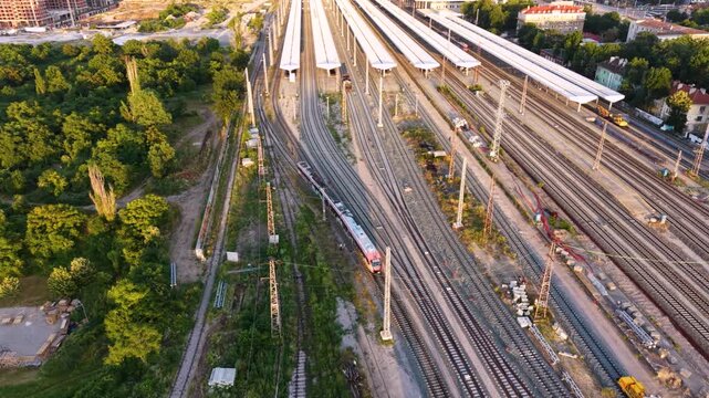Aerial sunset view of a large railway yard and train depot in Sofia, Bulgaria, with numerous passenger trains, parallel tracks, maintenance buildings and rail infrastructure extending through the city