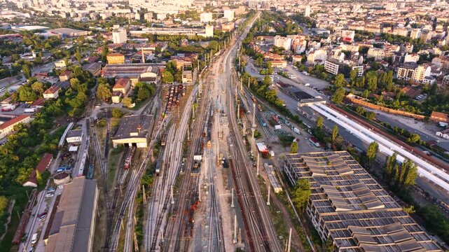 Aerial sunset view of a large railway yard and train depot in Sofia, Bulgaria, with numerous passenger trains, parallel tracks, maintenance buildings and rail infrastructure extending through the city