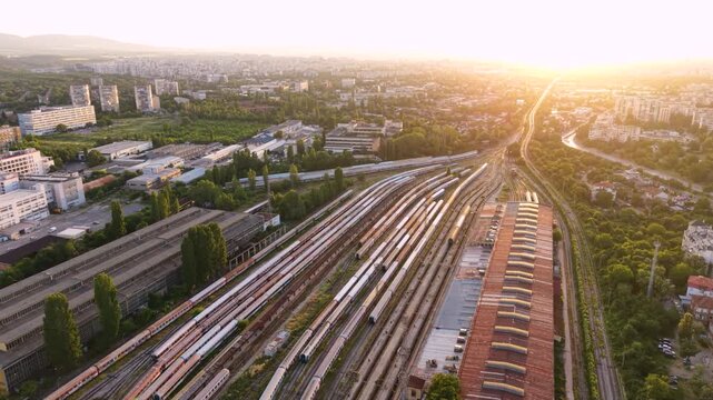 Aerial sunset view of a large railway yard and train depot in Sofia, Bulgaria, with numerous passenger trains, parallel tracks, maintenance buildings and rail infrastructure extending through the city