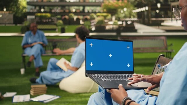 The medical student holds a laptop with a blue chroma key screen and uses the touchpad with one hand, creating an isolated editable display for computer screen replacement on campus.