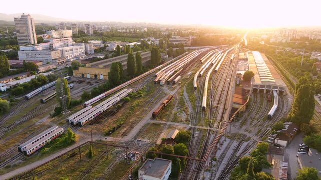 Aerial sunset view of a large railway yard and train depot in Sofia, Bulgaria, with numerous passenger trains, parallel tracks, maintenance buildings and rail infrastructure extending through the city