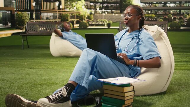 The portrait shows a Black medical student smiling and typing on a laptop while seated on a beanbag in a bright modern campus courtyard during outdoor medical school training, with study notes nearby.