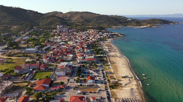 Aerial view of Toroni in Sithonia, Halkidiki, Greece, showing the coastal village beside a long sandy beach, clear turquoise Aegean Sea and rugged green hills in warm summer evening light.
