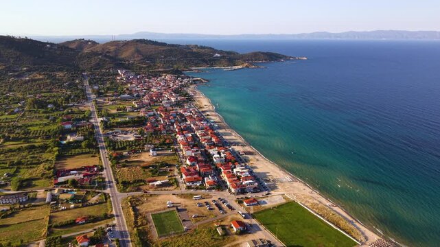 Aerial view of Toroni in Sithonia, Halkidiki, Greece, showing the coastal village beside a long sandy beach, clear turquoise Aegean Sea and rugged green hills in warm summer evening light.