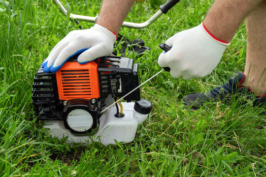 A person is pulling the starting cord of a lawn mower engine in a backyard. The grass is green, and the weather is clear. The individual wears gloves and is focused on the task