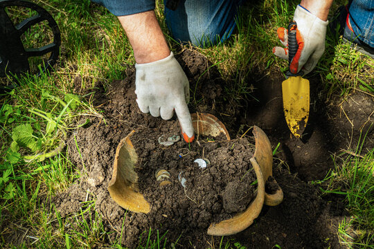 A treasure hunter is using a metal detector and digging in the soil of a garden. They are pointing at a pot that is half buried. The scene shows green grass and tools nearby