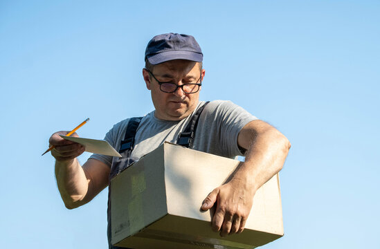 A delivery worker stands outside holding a cardboard box in one arm while looking at a note in his other hand. It is a bright day and he is focused on the details