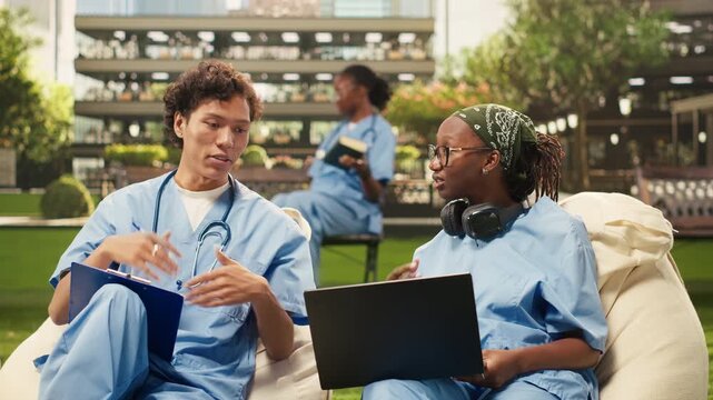Diverse med school peers in blue scrubs collaborate over laptop notes while seated on beanbags, studying coursework in a modern outdoor campus setting, during a group study session.