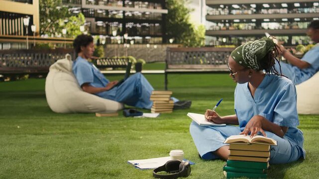 The medical student in blue scrubs writes in a notebook beside an open book and stacked books on sunny campus lawn, during outdoor learning.