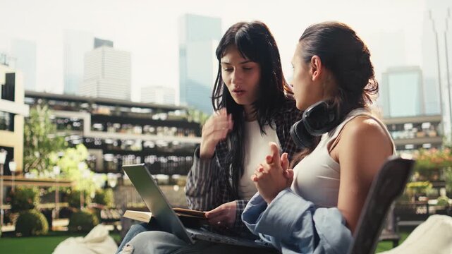 Team of university students sits on a bench with an open laptop and stacked books, smiling as they discuss project ideas in a modern campus courtyard, with classmates studying together.