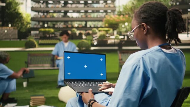 The laptop on the student's lap displays an isolated blue chroma key screen, on a university campus with peers in blue scrubs, with an editable mockup screen for replacement on the computer.
