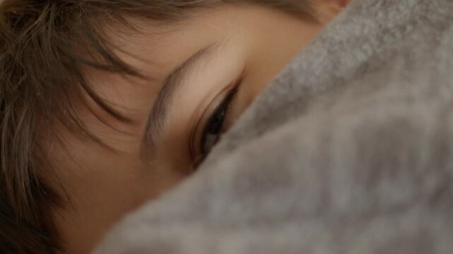 Soft gray blanket covers a smiling sleeping child. Child's eyes are closed, lips curved in gentle peace. Dark brown hair frames a calm, restful face. Warm, quiet moment of pure, sweet sleep
