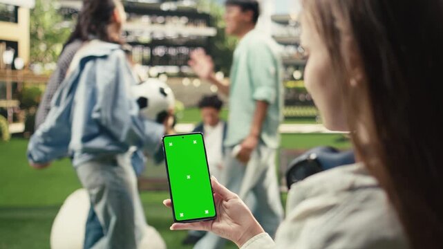 Vertical chroma key appears on an editable smartphone screen held by a young woman outdoors in a university campus study area, offering a green screen phone mockup for mobile screen replacement.