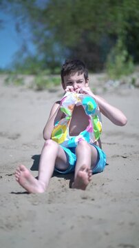 Shirtless child on the beach blows air into a bright multicolored inflatable swim ring under the sun, getting ready for aquatic play and leisure on holiday.