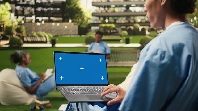 The laptop on the student's lap displays an isolated blue chroma key screen, on a university campus with peers in blue scrubs, with an editable mockup screen for replacement on the computer.