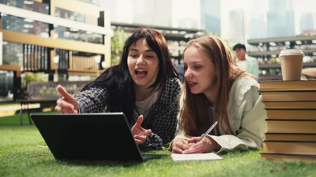 University students lie and lean over an open laptop and study materials on the grass, reviewing coursework in a textbook together during outdoor campus learning and peer tutoring for university life.