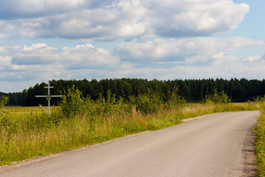 Tall Orthodox wayside cross standing by a quiet rural asphalt road with lush summer meadow, green forest edge under a blue sky with fluffy white clouds, Russia