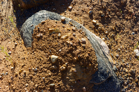 Large textured granite gneiss boulder covered with sand and small stones inside an open pit mine, displaying rugged geological patterns and raw earth tones under bright sunlight
