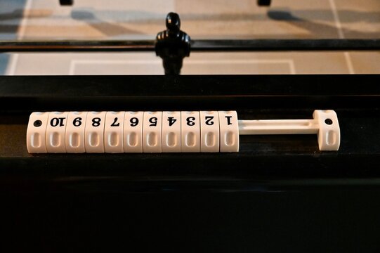 Close-up view of a mechanical score counter with white numbered cubes from 1 to 10 mounted on the edge of a foosball table, with a black player figure on a rod in the background.