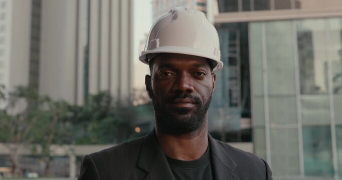Serious African American construction engineer in a white hardhat looks directly at the camera outside a modern office building in a confident professional portrait.
