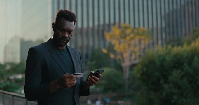 African American businessman enters payment details on a smartphone while holding a credit card outside the office, illustrating secure online shopping and fintech.