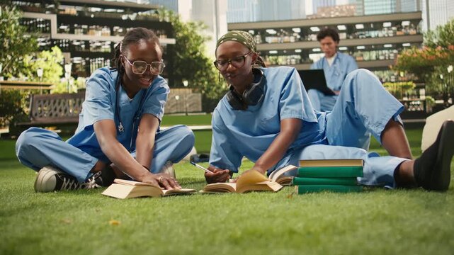 Medical school students in blue scrubs lean over open textbooks, studying together in a healthcare training program, with classmates collaborating beside stacked books on the lawn.