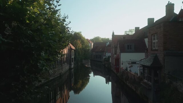 Narrow canal winds between historic brick houses and leafy trees in Bruges, Belgium, revealing a quiet residential corner of the medieval old town in morning light.