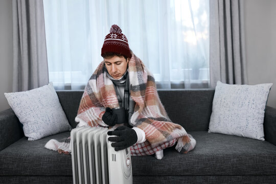Freezing man wrapped in a blanket near a heater during a cold snap. He faces a home heating problem, weak heating, and rising energy costs due to the winter energy crisis.