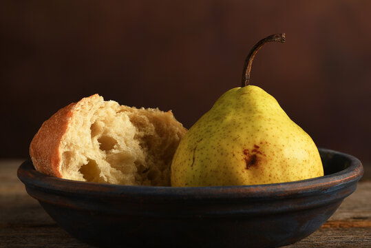 Pear and Bread Still Life. An Anjou Pear and a piece of broken bread on a plate and rustic wooden table with a mottled brown background.