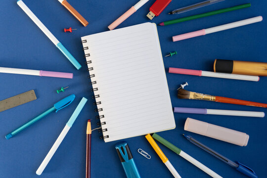 White spiral notebook surrounded by colorful stationery on blue surface