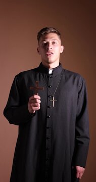 Priest in cassock with wooden cross praying on beige background