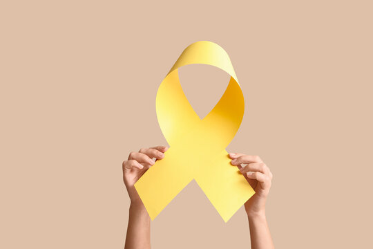 Hands holding a yellow awareness ribbon for cancer support and disease prevention on a neutral background, the ribbon represents hope, solidarity