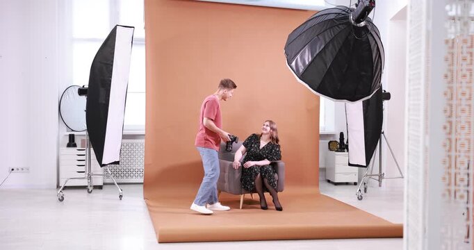 Young overweight woman in dress posing on armchair for photographer with camera against light brown background in studio. Plus size model