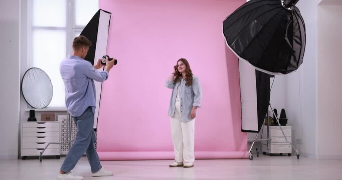 Plus size model posing for photographer with camera against pink background in studio. Beautiful young overweight woman