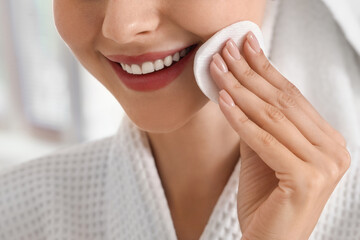 Smiling woman in a white bathrobe applying toner to her face with a cotton pad for Daily skincare routine © Pixel-Shot