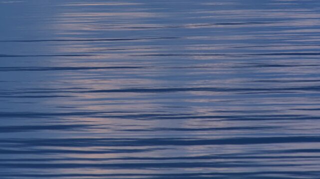 A close-up of the yellowish-orange moon's reflection on a calm blue sea in the early evening, where gentle waves and ripples create horizontal, parallel lines.