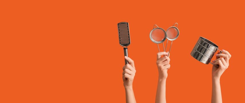 Hands holding kitchen utensils including a grater, two strainers, and a flour sifter on an orange background