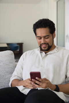 African American man sitting on sofa wearing henley and black watch holding red phone, smiling